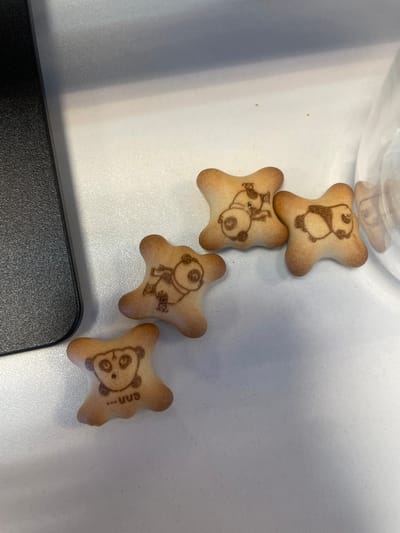 Four light brown, irregularly-shaped biscuits, each imprinted with a dark brown panda design, are arranged on a white, subtly textured surface. To the far left, a portion of a black, granular-textured object with a rounded corner, possibly an electronic device, is visible. On the right side of the frame, a clear, transparent object, likely a glass or container, is partially present, reflecting one of the biscuits. The biscuits depict different panda poses: one appears to be juggling while lying down, another is lying prone, and a third shows a panda head with wide eyes and the text "...uuu" below it. The scene suggests an indoor environment, likely a desk or tabletop. The specific location within Montigny-le-Bretonneux, France, cannot be identified from the visual information presented in the image.