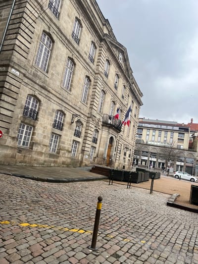 The image captures an exterior view of the Hôtel de Ville (City Hall) situated on Place Marouret in Le Puy-en-Velay, France, under an overcast sky. The prominent subject is a large, multi-story building constructed of light-colored stone blocks, exhibiting classical architectural elements including numerous rectangular windows with white frames and black wrought-iron balconies on the lower floors. Three French flags (blue, white, red vertical stripes) are suspended from a central second-story balcony. A street sign reading "PLACE MAROURET" is affixed to the building's left corner. The foreground features an uneven cobblestone street and sidewalk, with a yellow painted line visible on the cobblestones. A dark brown metal bollard with a yellow top stands in the immediate foreground. To the right of the main building, a small public area contains several dark wooden planter boxes and a couple of wooden benches, set on a light brown gravel or dirt surface. In the background, beyond this plaza, a more modern multi-story building with a light facade and numerous windows is visible, along with other urban structures and a single white car on a paved road. No discernible individuals are present in the scene.