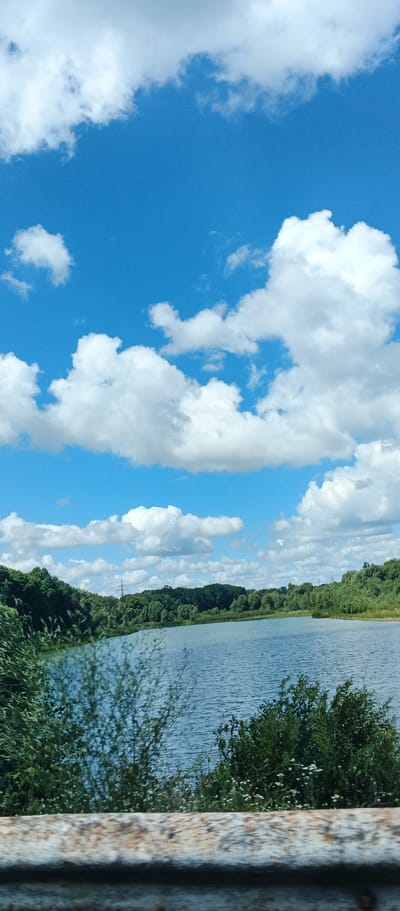 The image depicts a body of water, likely a lake or river, surrounded by lush green trees. The sky above is a vibrant blue, dotted with fluffy white clouds. In the foreground, a weathered, possibly metallic, railing or barrier runs horizontally across the bottom of the frame, slightly out of focus. The overall scene suggests a natural, tranquil environment. This particular view is situated in Kholodnianske, Ukraine, a location known for its natural landscapes and bodies of water.