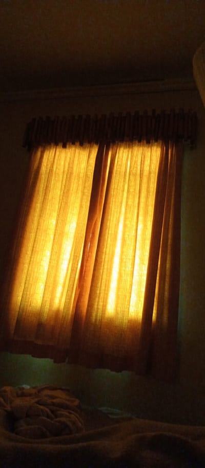 A dimly lit room reveals a window covered by thick, golden-yellow curtains. The curtains glow from the light streaming through, creating vertical columns of brightness. Above the curtains, a dark brown valance is visible. Below, a rumpled blanket covers what appears to be a bed. The room is generally dark, emphasizing the light shining through the window.