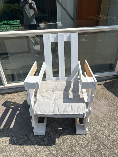 In Lisse, Netherlands, an outdoor scene features a white wooden chair with a gray cushion. The chair appears to be constructed from pallets and is positioned on a paved surface with visible brick patterns. Behind the chair is a large window, reflecting sunlight and showing a partial view of the interior. The reflection in the window includes a person holding a phone and text in Dutch, which translates to "ours VIDEO SURVEILLANCE. During opening hours, our store is not responsible for valuable items." The environment suggests it is a sunny day, with shadows cast on the ground.