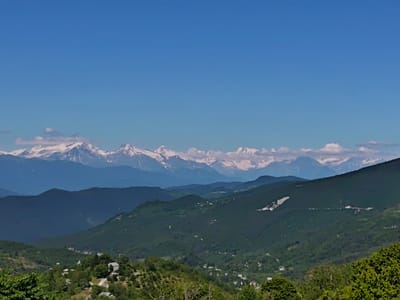 The image shows a panoramic view of a mountainous landscape in Ukeshi, Georgia. The foreground is dominated by lush green hills covered in trees and dotted with small houses. In the mid-ground, more rolling hills recede into the distance, also covered in dense vegetation. The background features a majestic mountain range with snow-capped peaks under a clear blue sky. The overall scene depicts a serene and natural environment, characteristic of a rural mountainous region.