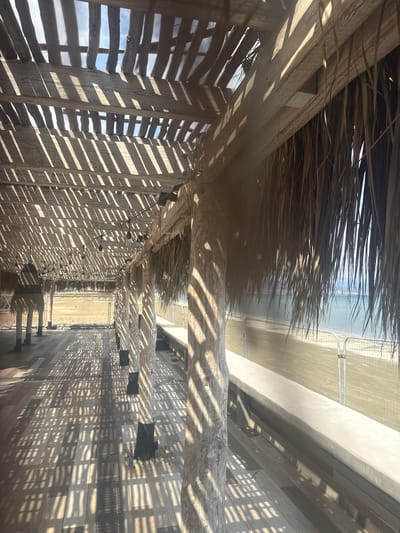 The scene depicts an empty, shaded outdoor walkway under a wooden pergola, situated in Unknown, Sovereign Base Areas of Akrotiri and Dhekelia. Prominent parallel striped shadows from the overhead wooden beams are cast across the multi-toned composite deck flooring. The pergola's roof consists of a lattice of wooden slats, with sections, particularly on the right side, incorporating dried palm fronds for additional shading. Multiple thick, light-colored columnar supports with dark bases anchor the structure. A string of clear bulb lights is strung horizontally between the beams, and a large, dark, conical-shaped woven lantern hangs from the pergola on the left. A linear metal grate runs down the center of the walkway. Along the right side, a low, wide concrete bench runs parallel to the path. Beyond this, a sandy beach is partially obscured by a temporary metal construction fence. The background reveals a calm, light blue-green sea under a clear sky. No individuals or active interactions are present.