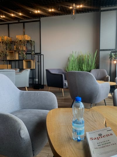 The image shows a cafe interior in Pyrzowice, Poland, located at the coordinates 50.471, 19.072. The scene is lit by overhead lights, and there are several chairs in the cafe, some gray and some blue. There are two wooden tables in the middle of the image, with a book and a bottle of water on one of them. The tables are made of wood. The room has a gray wall with a metal grid behind it. There are plants growing in the grid. The book on the table is titled *Sapiens: A Brief History of Humankind* by Yuval Noah Harari. The book appears to be in Russian. There is a bottle of water on the table next to the book. The bottle appears to be partially full of water. A person is not visible in the image.
