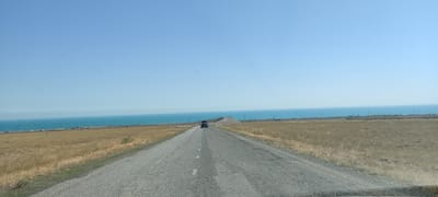 The image shows a view from a vehicle traveling on a two-lane asphalt road. The road is cracked and has white lines dividing the lanes. The vehicle, a dark SUV with luggage on its roof, is driving away from the camera towards a slight curve in the road. The landscape on either side of the road is a dry, grassy plain. In the distance, a wide expanse of turquoise water, likely a lake or sea, stretches to the horizon under a clear blue sky. Along the shoreline, there appear to be some structures and what might be a railway line or another road. The scene is consistent with the surroundings of Көктұма, Kazakhstan, a location known for its proximity to a large body of water and relatively flat, arid terrain.