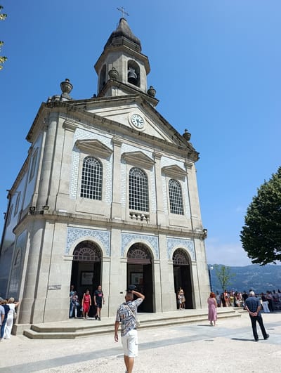 In Terras de Bouro, Portugal, a stone church with a prominent bell tower stands under a clear blue sky. The church facade is adorned with decorative blue and white tiles and features several arched windows. A clock face is visible on the tower. In front of the church, a paved plaza is bustling with activity. Several people are walking, standing, and gathered in small groups. A man in a patterned shirt and white shorts is walking away from the camera, facing the church entrance. Others are entering or exiting the building, and some are conversing on the plaza. The scene is bathed in bright sunlight, casting distinct shadows. In the background, a lush green tree stands to the right, and a hazy mountain range is visible in the distance.