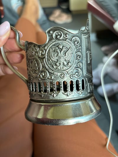 A person's hand with light pink fingernails holds a silver-toned metal cup holder. The cup holder is ornate, featuring an embossed double-headed eagle crest in the center, surrounded by swirling decorative patterns. Below the crest, there is a section with rectangular cutouts. The base of the cup holder is a wide, flared silver metal saucer. The background is blurred, showing what appears to be legs of a person wearing orange fabric and a white cord extending from the right side of the frame. The image is taken indoors, with a shallow depth of field. There is no indication of the city of Arsk, Russia, in this image.