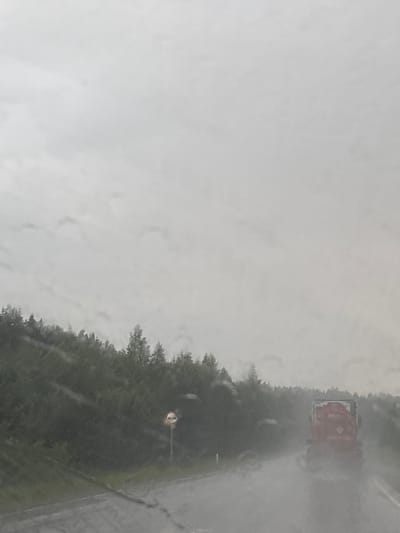 The image depicts a rain-soaked road with a forest lining the left side. A red tanker truck is driving on the wet road, creating spray. In the background, a circular traffic sign with two figures is visible on a pole. The sky is overcast and grey. The overall scene suggests poor weather conditions, likely on a highway outside of Bol'shoi Talmaz, Russia, given the presence of a sign that appears to be a Russian traffic sign. The view is from inside a vehicle, as indicated by water droplets on what seems to be a windshield.