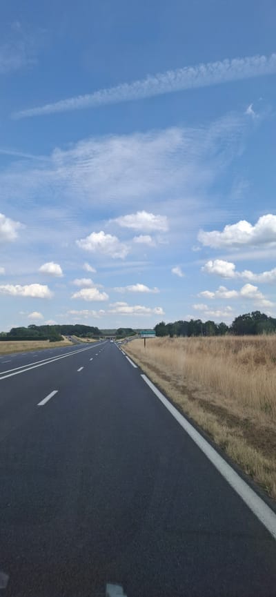 The media file captures a highway scene in Chailly-en-Bière, France. The perspective is from a vehicle traveling along the asphalt road, which is divided by white lines. To the left of the road, a grassy field stretches out, bordered by a line of trees. The sky above is a vibrant blue, dotted with fluffy white clouds and some wispy cirrus clouds. The time of day appears to be daytime, likely late morning or early afternoon, given the bright sunlight casting shadows. The weather seems pleasant with clear skies and scattered clouds. A green highway sign is visible in the distance, indicating directions and distances to various locations. The overall scene evokes a sense of travel and open road, with the natural landscape and blue sky suggesting a peaceful journey. No people are clearly visible in the media. The dominant colors are the dark grey of the asphalt, the golden brown of the dry grass, the green of the trees and sign, and the bright blue of the sky. The presence of the highway and the style of the sign suggest a location in France.