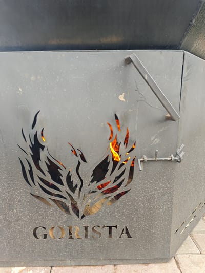 A medium shot captures a close-up of a dark grey, metal fire pit with a decorative cutout of a phoenix or bird of prey in flames. The fire pit has the word "GORISTA" cut out in large letters at its base. A metal latch is visible on the right side of the fire pit, securing a hinged door. The background is blurred, suggesting an outdoor setting with some paved ground visible at the bottom. The overall scene appears to be located in an outdoor urban environment, possibly a park or recreational area.