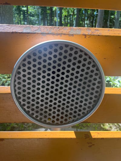A close-up, eye-level shot shows a circular metal grate with numerous small holes, positioned centrally on a weathered, orange-painted wooden surface. The grate appears to be part of an outdoor fixture, possibly a trash receptacle or ventilation cover. The wooden surface is slightly distressed, with visible paint chips and grain. Behind the grate, through the wooden slats and the holes in the grate, a blurred background of green trees and foliage is visible, suggesting an outdoor, possibly park-like setting. The sunlight casts strong shadows, highlighting the texture of the wood and the perforations of the grate. There are no people or animals visible in the image. The setting is likely Guryevsk, Russia, based on the context provided.