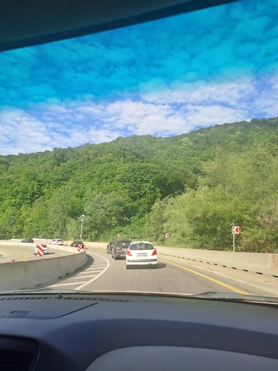 The image depicts a highway scene in the vicinity of Moldavanovka, Russia. The perspective is from inside a vehicle, looking forward through the windshield. A bright blue sky with scattered white clouds dominates the upper portion of the frame. A dense, green forest covers a hillside to the right of the highway, extending upwards and towards the left. The highway itself curves to the left, with concrete barriers lining the sides. Several vehicles are visible on the road, including a white sedan in the foreground, a black SUV, and other cars further ahead. Road signs, including directional signs indicating curves and possibly speed limits, are present along the roadside. The overall impression is of travel on a scenic, wooded highway.