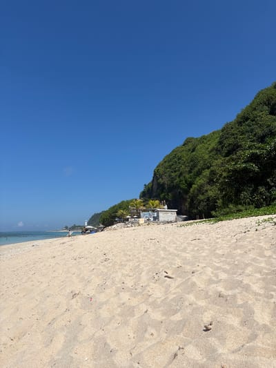 The image captures a beach scene in Kutuh, Indonesia, presenting a clear blue sky that occupies a substantial portion of the frame. To the right, a sandy beach slopes toward a strip of water along the bottom edge of the image.
Adjacent to the beach is a densely vegetated cliff face. Towards the middle ground, structures are nestled along the cliff's edge, suggesting a resort or private dwellings. Several people are visible along the shoreline. The sand texture indicates footprints and disturbances.