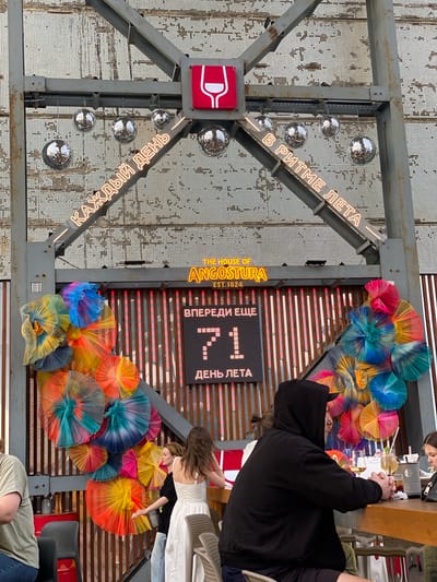 The image depicts an outdoor event space, possibly a bar or restaurant, decorated with a large metal structure adorned with disco balls and neon signs. The neon signs in Cyrillic text read "КАЖДЫЙ ДЕНЬ" (Every day) and "В РИТМЕ ЛЕТА" (In the rhythm of summer). Below this, a sign identifies "THE HOUSE OF ANGOSTURA EST. 1824". A digital display shows the number "71" with the text "ВПЕРЕДИ ЕЩЕ" (Still ahead) above it and "ДЕНЬ ЛЕТА" (Day of summer) below it. The area is further decorated with vibrant, colorful paper fans. Several people are present, some seated at a bar, others standing. One person in the foreground is wearing a black hooded sweatshirt and is seated at the bar. A woman in a white dress is standing near the decorative fans. The overall atmosphere suggests a celebratory or festive occasion. There is no indication of the specific location being Valdepeñas, Spain; the image itself does not provide any geographical markers to confirm this.