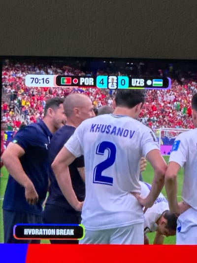 The image shows a screen displaying a football match. The score indicates Portugal (POR) is leading Uzbekistan (UZB) 4-0, with the game at 70:16. A "HYDRATION BREAK" notification is visible on the right side of the screen.
The foreground features several people, likely football players, in a huddle. One player in a white jersey has "KHUSANOV" and the number "2" on his back. Other players are wearing dark blue jerseys. The background shows a crowded stadium filled with spectators. The setting is a football match, indicated by the score overlay and the presence of players and a stadium. The lighting suggests it is daytime. The mood appears to be one of camaraderie or celebration among the players. The image is captured from a slightly elevated, oblique angle, focusing on the players and the screen content. The red and blue vertical bars on the right edge of the screen are likely part of the display interface or a border.