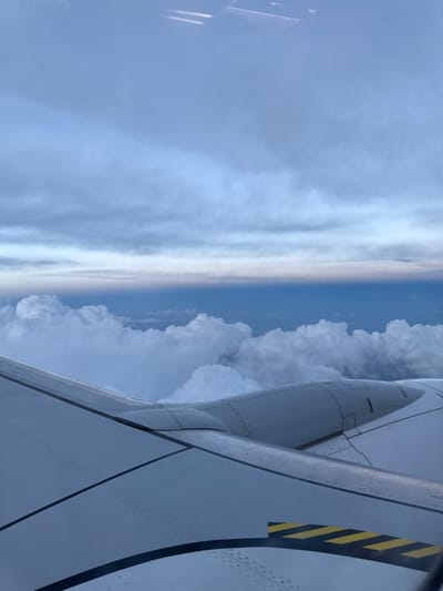 From an airplane window, a wing partially obstructs the view of a sky divided by cloud formations. To the left, a broad expanse of hazy, grayish-white clouds extends to the horizon. On the right, a more defined cluster of white cumulus clouds rises against a blue sky. The airplane wing is predominantly white with visible paneling and rivets, featuring a black and yellow striped warning marking near the edge.