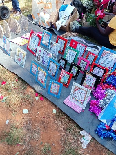 A black tarp displays an array of framed graduation certificates and cards. The certificates are decorated with colorful borders and celebratory messages. Flowers, graduation sashes, and tinsel garlands accompany the certificates. A person sits nearby arranging roses. The ground around the tarp is grassy, with scattered rose petals. A few shopping carts and bags are visible.