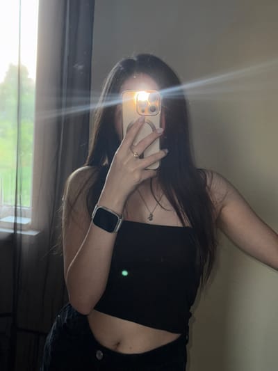 The media file shows a person taking a mirror selfie, rotated 90 degrees counter-clockwise. The subject is a person with long, dark hair, wearing a black crop top and dark pants. They are holding a smartphone in their right hand, with a ring on their left hand. A smartwatch is visible on their left wrist. The reflection in the mirror shows the person's arm and shoulder. There is a bright light glare across the image, obscuring the person's face. The background appears to be a wall, with a window or curtain visible at the bottom, suggesting an indoor setting. The lighting indicates it could be daytime, possibly with sunlight streaming in. No text is visible in the image. The location context is Horenychi, Ukraine.