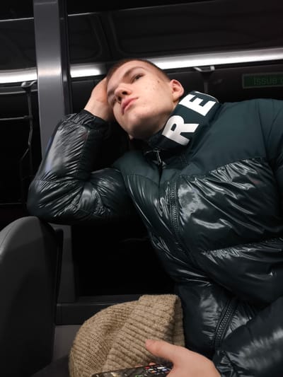 A young man with short-cropped hair rests his head on his hand while seated. He wears a shiny, dark green puffer jacket with "RE" emblazoned on the white collar. A beige knit cap rests in his lap, and he holds a remote control. The setting appears to be the interior of a train or bus, with dark, reflective surfaces visible in the background and a window above. The lighting is dim and subdued.