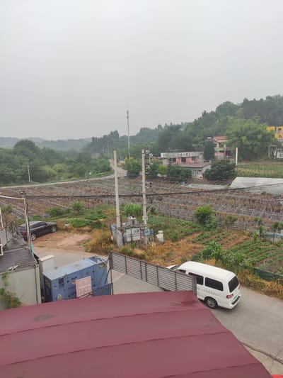 This image depicts a rural or semi-rural landscape, likely on the outskirts of Jianyang City, China, on an overcast day. In the foreground, a dark-colored car is parked on a dirt and gravel area. To its left, a large blue generator unit with "BaiFa" and "Power" branding is situated next to a utilitarian building. A red-tiled roof occupies the bottom of the frame.

Beyond the immediate foreground, a series of agricultural greenhouses, characterized by their arched metal frames and plastic coverings, dominate the mid-ground. Rows of crops, including young corn plants, are visible in the open fields adjacent to the greenhouses. A paved road winds through the scene, and a white van is traveling on it, heading away from the camera.

In the background, rolling hills covered in lush green trees rise, interspersed with scattered buildings and houses, some of which appear to be residential. A prominent telecommunications tower stands on one of the distant hills. Power lines and utility poles are visible throughout the landscape, indicating the presence of infrastructure. The overall atmosphere is one of a working agricultural community under a muted sky.