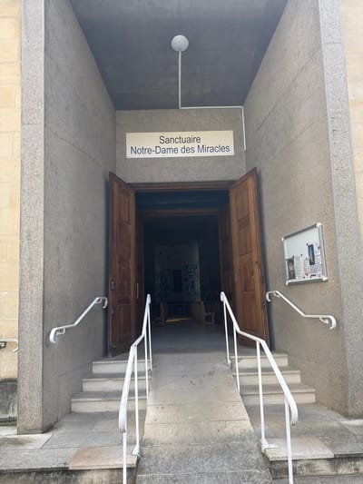 The image displays the entrance to the Sanctuaire Notre-Dame des Miracles in Orléans, France, viewed from an exterior, slightly low vantage point. The entrance is recessed within a modern-style facade of textured grey concrete, flanked on the far left by an older, lighter-colored stone wall. A white, circular ceiling light fixture is centered above the entry. Two tall, dark brown wooden double doors are open inwards, revealing a dimly lit interior with visible benches or pews and abstract, possibly stained-glass artwork in the background.
Access to the doors is provided by a central concrete ramp flanked by two sets of concrete steps. White metal handrails, showing some wear, ascend on both sides, accompanying both the ramp and the adjacent steps. On the right concrete wall, a silver-framed notice board displays several informational papers and images. The scene is devoid of people and illuminated by natural daylight.
