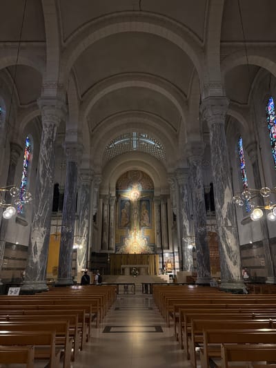 This is a full shot of the interior of a large church, likely a basilica, in Annecy, France. The perspective is from the nave, looking towards the altar at the far end. The architecture is grand, with soaring vaulted ceilings supported by massive marble columns. Sunlight streams through tall stained-glass windows on either side, casting colorful patterns on the floor. Rows of empty wooden pews fill the nave, with a central aisle leading to the altar. The altar area is adorned with a large mosaic depicting a religious scene, likely Christ on the cross. Two figures, a man and a woman, stand near the altar, appearing to be attending a service or prayer. The overall atmosphere is one of solemnity and reverence.