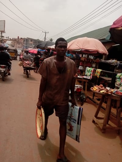 The image depicts a bustling street scene in Umudioka, Nigeria, under an overcast sky. In the foreground, a dark-skinned adult male walks directly towards the viewer, wearing a dirty brown V-neck shirt, dark shorts, and flip-flops. He carries a white and red oval tray in his left hand and a blue and white striped "SUPREME SEMOLINA" branded bag in his right hand. To his right, a market stall is visible with a large pink umbrella providing shade over tables displaying various packaged goods, including yellow and red rectangular blocks on a metal tray. To his left, further down the reddish-brown unpaved street, a man in a light-colored shirt pushes a wheelbarrow laden with items. A person on a motorcycle is partially visible to the far left. In the mid-ground, other market umbrellas (including a black one) and indistinct figures indicate further market activity. The background features multi-story buildings and numerous overhead utility lines crisscrossing the sky, supported by utility poles.