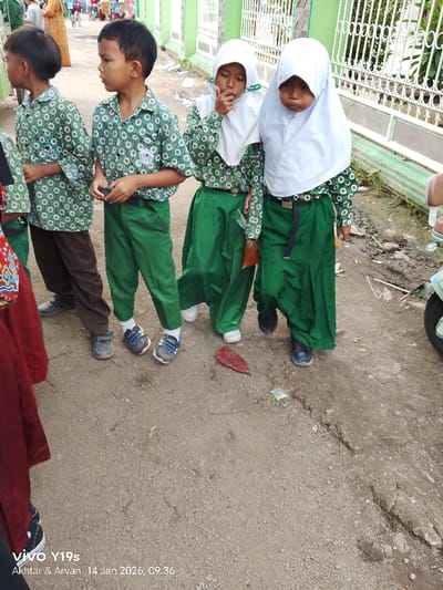 A group of elementary-aged children are standing on a dirt path near a white metal fence. The children are dressed in matching green and white school uniforms. The girls wear white hijabs. The boys wear short-sleeved button-down shirts. The lower half of the boys' uniform consists of green pants. The girls are wearing green skirts. A few people can be seen in the background.