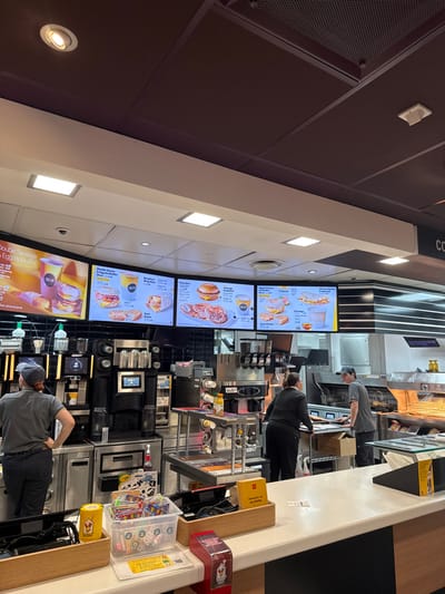 This is an interior view of a McDonald's fast-food restaurant, located in East Hampshire, United Kingdom. The scene depicts the service counter and kitchen area from a customer's perspective. The ceiling is segmented, featuring dark brown panels and white suspended sections with square and circular recessed lighting fixtures. Five illuminated digital menu boards display breakfast items, including "Double Sausage & Egg McMuffin," "Pancakes," and "Cheesy Bacon Flatbread," positioned above a black-tiled backsplash. Three uniformed employees are visible behind the counter. On the left, an employee in a dark grey shirt and cap stands with their back to the viewer, hands on hips. In the central right, a second employee in a dark long-sleeved uniform attends to equipment. A third employee, wearing a grey shirt and cap, is further right near hot food service equipment, facing the central employee. The foreground counter is white with a dark base, displaying a yellow McDonald's-branded cup, a clear plastic container filled with small toys, a yellow laminated document, a red dispenser, and a wooden box labeled "MCDONALD'S ALLERGEN INFO." The background features extensive stainless steel commercial kitchen equipment, including multiple beverage dispensers, fryers, and warming units.