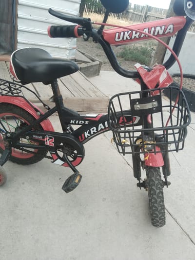 A red and black children's bicycle is parked on a concrete surface. The bicycle has a black seat, red fenders, and a black basket on the front. The word "UKRAINA" is printed in large white letters on a red bar across the handlebars. The bicycle also has the word "KIDS" printed on the frame. A training wheel is visible on the left side of the rear wheel. The background shows a wooden fence and some vegetation. The location is Ақадыр, Kazakhstan.