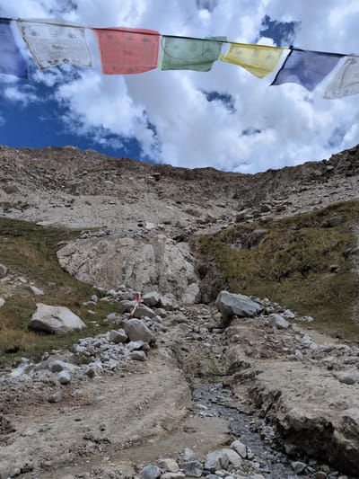 The image depicts a rugged, rocky mountain slope in Panamik, India, under a cloudy sky. Prayer flags, displaying various colors including blue, white, red, green, and yellow, are strung across the upper portion of the frame, fluttering in the wind. The mountainside is covered in loose stones, scree, and patches of dry grass, suggesting a high-altitude, arid environment. A narrow, shallow stream or dry creek bed winds its way down the slope, marked by scattered rocks and a faint track of disturbed earth. In the middle ground, a small cluster of prayer flags are tied to a rock face, adding a splash of color against the natural terrain. The overall impression is one of a remote, windswept landscape characteristic of mountainous regions.