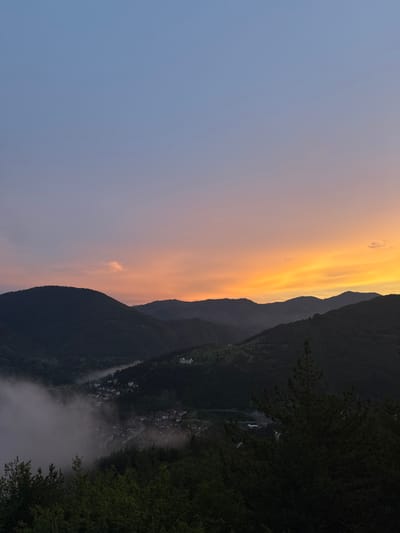 The image depicts a panoramic view of the city of Strazha, Bulgaria, at dusk. The sky transitions from a soft blue at the top to a vibrant orange and yellow glow near the horizon, indicating the setting sun. Silhouetted against the colorful sky are several layers of rolling hills covered in dark green trees. In the valley below, the lights of Strazha are visible, partially obscured by a layer of mist that adds a dramatic effect to the scene. The mist appears to be settled in the lower parts of the valley, surrounding the buildings of the city. In the foreground, the dark outlines of trees are visible, framing the view. A large, light-colored building, possibly a hotel or a monastery, is situated on one of the higher slopes overlooking the city. The overall impression is one of a tranquil and picturesque mountain town as day turns into night.