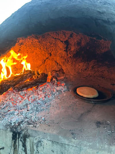 The image provides an internal view of a rustic, wood-fired oven or fire pit, focusing on the active combustion and an item being cooked. The perspective is angled downwards, looking into the heat source.

On the left side of the frame, the inner wall of the oven, possibly constructed from clay or earth, glows with a warm, intense orange and reddish hue, illuminated by the fire. This surface is rough, textured, and appears to be absorbing and radiating heat.

At the bottom center, a vibrant fire is burning intensely. Bright orange and yellow flames dance around several dark, partially consumed logs of wood, which are providing the heat for the oven.

To the right of the active flames, a bed of glowing red embers is visible, intermingled with significant amounts of light grey ash. This area indicates where wood has already burned down, still radiating considerable heat.

In the upper right section of the image, a circular, flat, light-colored object, resembling a piece of unbaked or partially baked bread (possibly flatbread or a similar dough product), rests inside a dark, round, shallow pan or dish. This pan is placed directly onto a flat, dark grey surface within the oven, which appears to be made of stone, concrete, or a similarly sturdy, fire-resistant material. This surface shows signs of charring and soot buildup.

The image captures a moment of traditional cooking, highlighting the raw elements of fire, ash, and natural oven materials. The lighting is dominated by the warm glow of the fire, creating strong contrasts and a sense of intense heat. There is no visible text in the image.