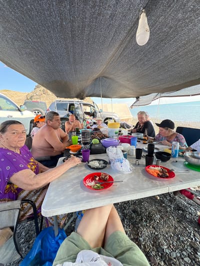 A group of people are gathered around a table for a meal outdoors. The scene appears to be on a rocky shoreline, with the sea visible in the background. Several vehicles are parked nearby, suggesting a camping or picnic outing. A large tarp provides shade over the gathering. Food and drinks are spread across the table, and some individuals are actively eating. The overall atmosphere is casual and social. The location is identified as the city of Karashygan, Kazakhstan.