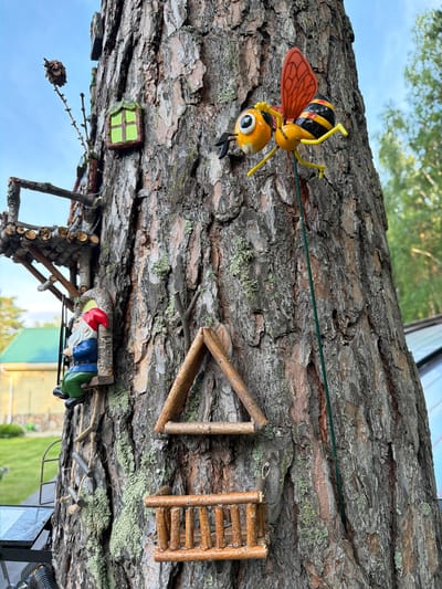 This media file captures a whimsical, miniature scene set on the rough bark of a tree. The primary subjects are several small, decorative figures and constructed elements, creating a fairy garden or gnome village.
Dominating the upper part of the frame is a toy bee with vibrant yellow and black stripes, orange wings, and large, expressive eyes. It is suspended by a thin green wire, suggesting movement or flight. Below the bee, a small, green-paned window is embedded in the bark, resembling a tiny house feature. Further down, a gnome figure with a red hat, blue jacket, and green pants is depicted sitting on a miniature log bench, seemingly relaxed. Adjacent to the gnome is a small ladder made of twigs, and further up the tree trunk, another twig ladder and a triangular structure made of twigs are visible. A pinecone dangles from a small branch on the left side of the image, adding to the naturalistic elements of the scene. The background shows a blurry view of green foliage, a bright blue sky, and hints of a house with a green roof, suggesting an outdoor setting.
The scene appears to be set during the daytime, with bright natural light illuminating the details. The focus on miniature, handcrafted elements and the natural setting of the tree bark implies a creative and playful display, likely in a garden or backyard. There is no visible text in the media file. The overall mood is charming and imaginative.