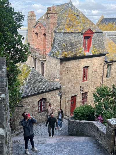 This image depicts a view from an elevated position looking down a stone stairway towards a group of tourists at Le Mont-Saint-Michel, France. The architecture is characterized by stone buildings with steeply pitched, weathered slate roofs, some adorned with yellow lichen. Several red-framed windows are visible, adding a contrasting color to the stone facades. A prominent brick structure with arched windows and a decorative balcony is visible in the background.

In the foreground, a Black man wearing sunglasses, a grey jacket, black pants, and white sneakers is standing on the stairs, holding a red-cased smartphone up to take a photo. To his right and slightly further down the stairs, a man wearing a dark green jacket and dark pants is walking. Behind him, a woman in a dark jacket and jeans is walking down the stairs. Further up the stairs, a man wearing a light brown jacket is standing and looking towards the right. The stairway is paved with cobblestones, and a stone wall with vegetation runs along the right side of the stairs. Lush green trees and bushes are interspersed with the buildings. The sky is overcast with scattered clouds.