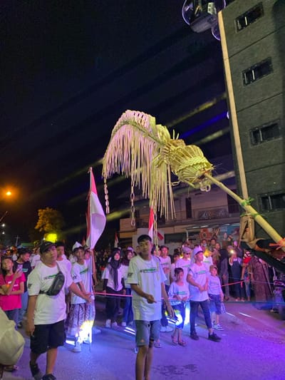 The image depicts a nighttime procession taking place on a paved street in Kedungbanteng, Indonesia. A large, ornate cultural effigy constructed from woven bamboo and palm fronds, featuring an arched canopy and a central spherical element, is prominently displayed, supported by a thick bamboo pole extending from the right foreground.

Numerous individuals, primarily young adults and children, are participating in or observing the event. In the foreground and mid-ground, a procession of people moves from left to right. Many wear white t-shirts, some bearing green text or logos. Several participants hold vertical Indonesian flags (red and white). A young girl in a pink shirt is visible on the far left, while a person in dark, potentially traditional attire, is partially visible on the far right. Spectators line the sides of the street in the background.

The scene is illuminated by artificial lights, casting a distinct purple and blue hue on the ground and lower sections of the participants. Streaks of light are visible against the dark night sky. A streetlamp on the left emits a yellow glow. Multi-story buildings with illuminated windows are visible in the background. A tall, dark line array speaker system is positioned on the upper right side of the frame, angled towards the procession.