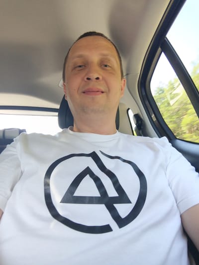 A middle-aged man with a shaved head and light skin is taking a selfie in the back seat of a car. He is wearing a white t-shirt with a large black circular logo containing an inverted triangle. The man is smiling slightly and looking at the camera. The interior of the car is visible, including the headliner and the passenger-side window, through which trees and foliage can be seen passing by, suggesting the car is in motion. The lighting indicates it is daytime. The location is described as Астроміцкі сельскі Савет, Belarus.