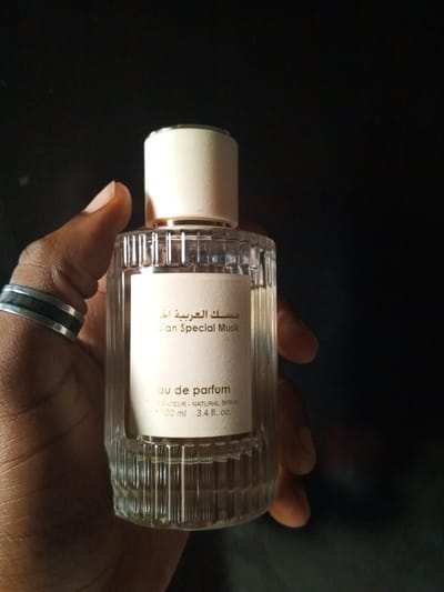 A person's hand wearing a ring is holding a glass bottle of "Arabian Special Musk" perfume against a dark background. The bottle has a ribbed glass base and a cream-colored cap. The label on the bottle is light-colored and features text in both Arabic and English. The image is a close-up shot, focusing on the bottle and the hand. The setting is not indicative of any specific location within the city of Ashata, Nigeria, or any other city.