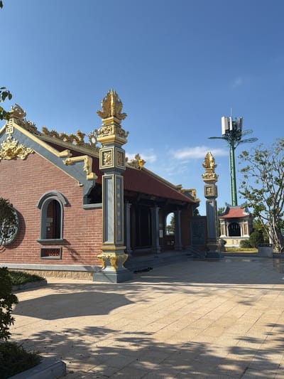 The image depicts a Buddhist temple in Diên Lạc, Vietnam. The temple is constructed of red brick, with a traditional red-tiled roof adorned with golden dragon motifs. A decorative post stands adjacent to the temple, also featuring golden embellishments. In the background, a modern cellular tower is visible, contrasting with the traditional architecture. The scene is bathed in sunlight, casting shadows on the paved courtyard. The sky is clear and blue. No people are visible in the frame.