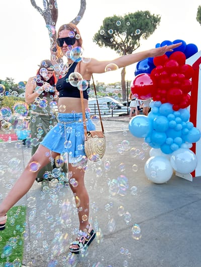 The image depicts a woman in a black crop top and a blue denim skirt, adorned with a gold chain belt, standing amidst a flurry of soap bubbles. She is wearing oversized black sunglasses and multi-colored sandals. Her left arm is extended, and her right arm is bent, holding a straw purse. She is positioned in the foreground, with another woman in a green outfit visible behind her. The background features trees, a street with parked cars, and a balloon arch composed of red, blue, and white balloons. The scene appears to be an outdoor event, possibly a celebration or party, in the city of Sant Vicenç De Montalt, Spain, indicated by the context of the request.