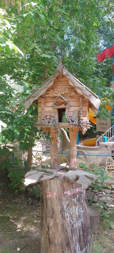 A whimsical, hand-carved wooden birdhouse, designed to resemble a miniature fairytale dwelling, stands as the central subject of this outdoor scene. The birdhouse is elevated on two intricately carved wooden legs that are anchored to a large tree stump. The stump itself is adorned with faded chalk drawings, suggesting it may have served as a canvas for children's art. The birdhouse features a detailed, gabled roof with decorative carvings, a small open doorway, and tiny wooden fences. Surrounding the birdhouse, lush green foliage from trees and bushes creates a natural, verdant backdrop. In the background, elements of a playground are visible, including colorful structures, slides, and railings, indicating the setting is likely a park or recreational area within the city of Berdsk municipality, Russia. The lighting suggests it is daytime, with sunlight filtering through the leaves. There are no people present in the image.