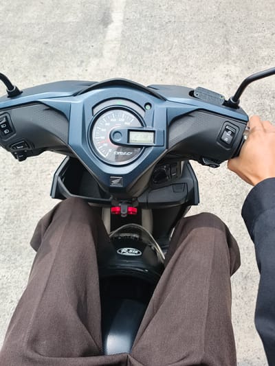 The image presents a first-person, overhead view from the perspective of someone seated on a dark blue and black Honda Beat scooter. The rider's legs, covered in brown trousers, are visible, as is their right hand gripping the throttle on the handlebar. A black motorcycle helmet, bearing a white "R-SIX" logo, rests on the scooter's floorboard between the rider's legs.
The scooter's dashboard is central, featuring an analog speedometer marked in "km/h" up to 140, with speed increments at 20, 40, 60, 80, 100, and 120. A digital display shows "2138.7", likely an odometer reading. Other visible texts and symbols on the dashboard include "HONDA", "PIGM-FI FUEL INJECTION", an "ECO" indicator light, headlamp symbols, and a horn symbol. The right handlebar features an engine start/stop switch labeled "A" and "OFF", and a brake fluid reservoir with warnings in English ("USE ONLY DOT3 OR DOT4 BRAKE FLUID"), Japanese ("ラスクブレーキ専用油をご使用下さい。"), and French ("LIQUIDE DE FREIN DOT3 OU DOT4 SEULEMENT"). An ignition switch with the word "SEAT" is also visible.
The scooter is positioned on a light gray, textured paved surface, likely concrete or asphalt, suggesting an outdoor setting during daylight hours. The background is out of focus, highlighting the immediate foreground. The scene depicts a common mode of transport, consistent with the provided location context of Gadog, Indonesia.