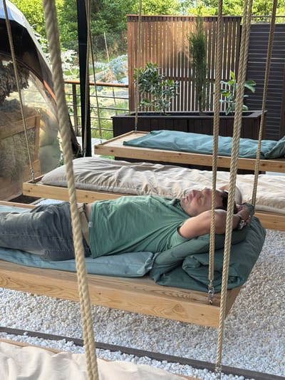 A man is lying on a suspended wooden platform bed, outdoors in what appears to be a glamping or resort setting. He is wearing a green t-shirt and blue jeans, with his head resting on a pillow and his hands behind his head. The bed is covered with a blue duvet and a beige sheet. The platform is suspended by thick ropes attached to overhead structures. In the background, another similar platform bed with a green duvet is visible. The area is covered with white gravel, and there is a wooden fence and lush green trees in the distance. The lighting suggests it is daytime, likely morning or afternoon. The location is specified as Nizhnyaya Shilovka, Russia.