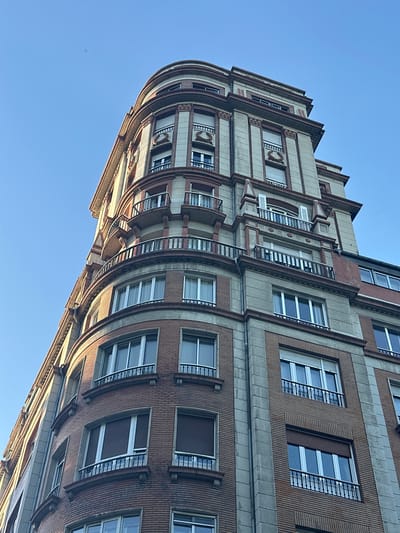 The image shows a low-angle, upward view of a multi-story building with a curved facade against a clear blue sky. The building is constructed with brick and light-colored stone elements, featuring decorative architectural details such as columns, pilasters, and balconies with wrought-iron railings. Many windows are visible, some with internal blinds drawn, reflecting the bright sky. The architectural style suggests it is an older building, possibly from the early to mid-20th century. This building is located in Oviedo, Spain, specifically on Calle Uría, a prominent street known for its elegant architecture. There are no people visible in the image.