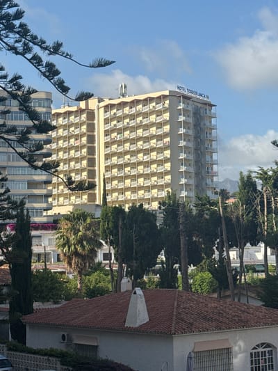 The image depicts a view of the Hotel Torrenblanca in Fuengirola, Spain, under a partly cloudy blue sky. The hotel is a large, multi-story building with a yellow facade, numerous balconies with white railings, and a spiral staircase on one side. In the foreground, a large pine tree branch partially obstructs the view of the hotel and another taller, modern building. Several other buildings, including a foreground house with a tiled roof and an air conditioning unit, are visible, along with various trees and lush greenery. In the background, mountains can be seen. The scene appears to be captured during daylight.