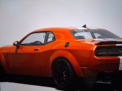 An orange Dodge Challenger with white racing stripes is parked on a white surface, possibly a display floor or studio. The car is viewed from the rear-quarter angle, showcasing its wide fenders, black wheels, and prominent rear spoiler. The background is a plain, light gray, indicating an indoor setting. There are no people or other objects visible in the scene. The image does not contain any elements that would suggest a location in the city of Argun, Russia.
