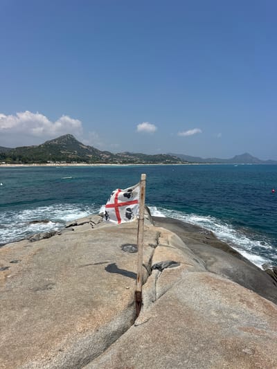 This is a photograph taken on a bright, sunny day in Castiadas, Italy, likely in the Costa Rei area, known for its beautiful beaches. In the foreground, a flag with the emblem of Sardinia, also known as the "Four Moors," is planted on a rough, granite rock formation. The flag is white with a red cross and four black heads with bandanas. The rock formation juts out into the clear, blue Mediterranean Sea, with gentle waves lapping against it. In the mid-ground, the sea stretches towards a coastline lined with a sandy beach dotted with sunbathers and umbrellas. Beyond the beach, rolling green hills rise, forming the backdrop of the scene, with a prominent, somewhat rugged mountain peak visible. The sky is a vibrant, clear blue with a few scattered white clouds. The overall atmosphere is one of a serene, picturesque coastal landscape. There are no visible people on the rocky foreground or engaged in any specific actions.