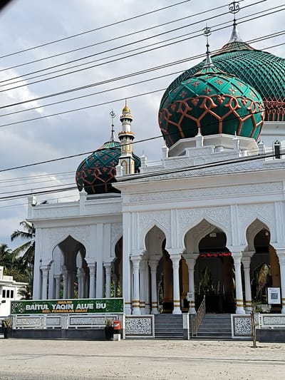 The image shows the Baitul Yaqin Alue Bu mosque in Aluebutuha, Indonesia, in the Peureulak Barat district. The mosque is a white building with arched entrances and columns, topped with distinctive green and orange-patterned domes. A minaret is visible beside one of the domes. A green sign in front of the building displays the mosque's name in English: "BAITUL YAQIN ALUE BU, KEC. PEUREULAK BARAT". A set of stairs leads up to the mosque entrance. Power lines are visible against the cloudy sky in the background.