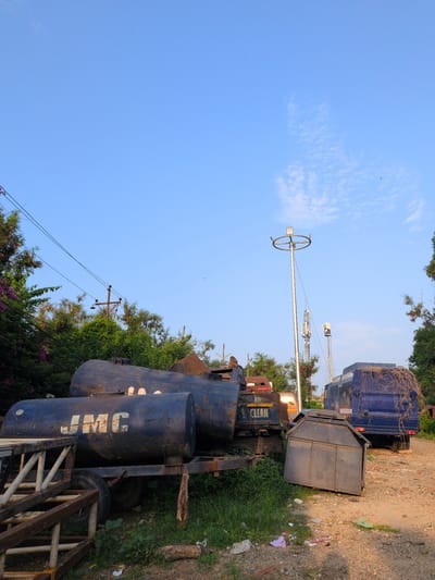 In Jammu, India, this image depicts an outdoor scene with a clear blue sky. In the foreground, a rusty, blue trailer holds two large cylindrical tanks. The front tank displays the white lettering "JMC". To the right, a blue garbage truck is parked on a dirt road. A small, dark metal bin is positioned on the ground in front of the garbage truck. The background features dense green foliage, a utility pole with electrical wires, and a tall lamppost with a circular fixture at the top. The ground is covered with dirt, grass, and scattered pieces of trash.